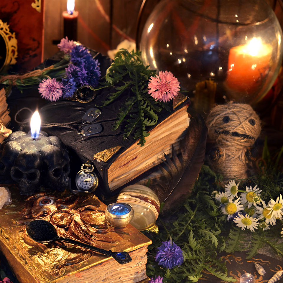 Various hoodoo objects on a table including a book, a doll, candles, and flowers