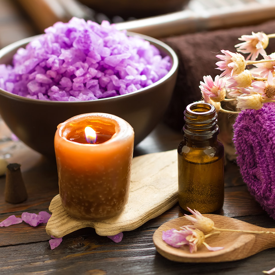 Aromatherapy products, including a candle and oil. Flowers, a bowl, and wooden spatulas are also visible in the image