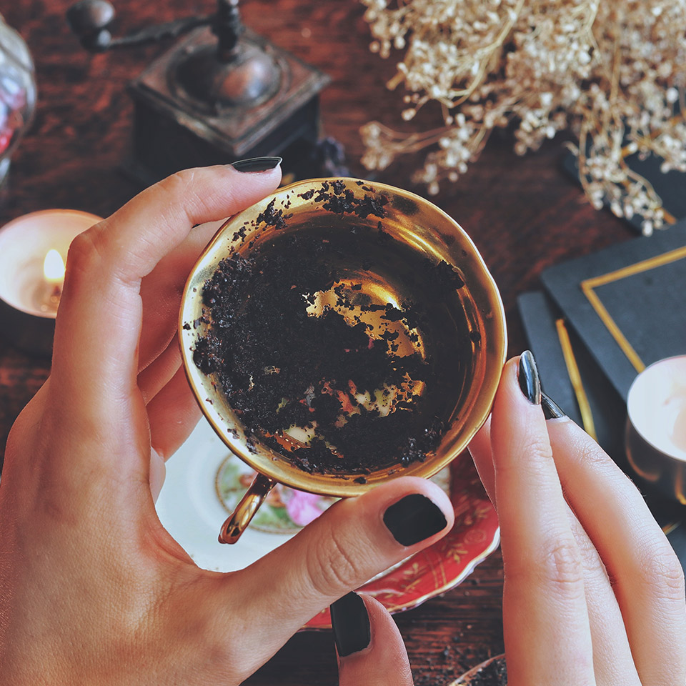 Hands holding a golden teacup for tea leaf tasseography. Black tea residue can be seen in the bottom of the cup.